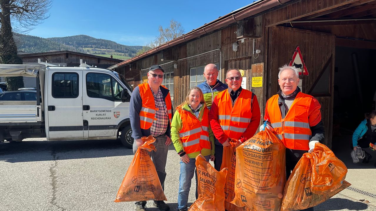 Five people in orange vests holding orange bags in front of a building with a white van in the background.