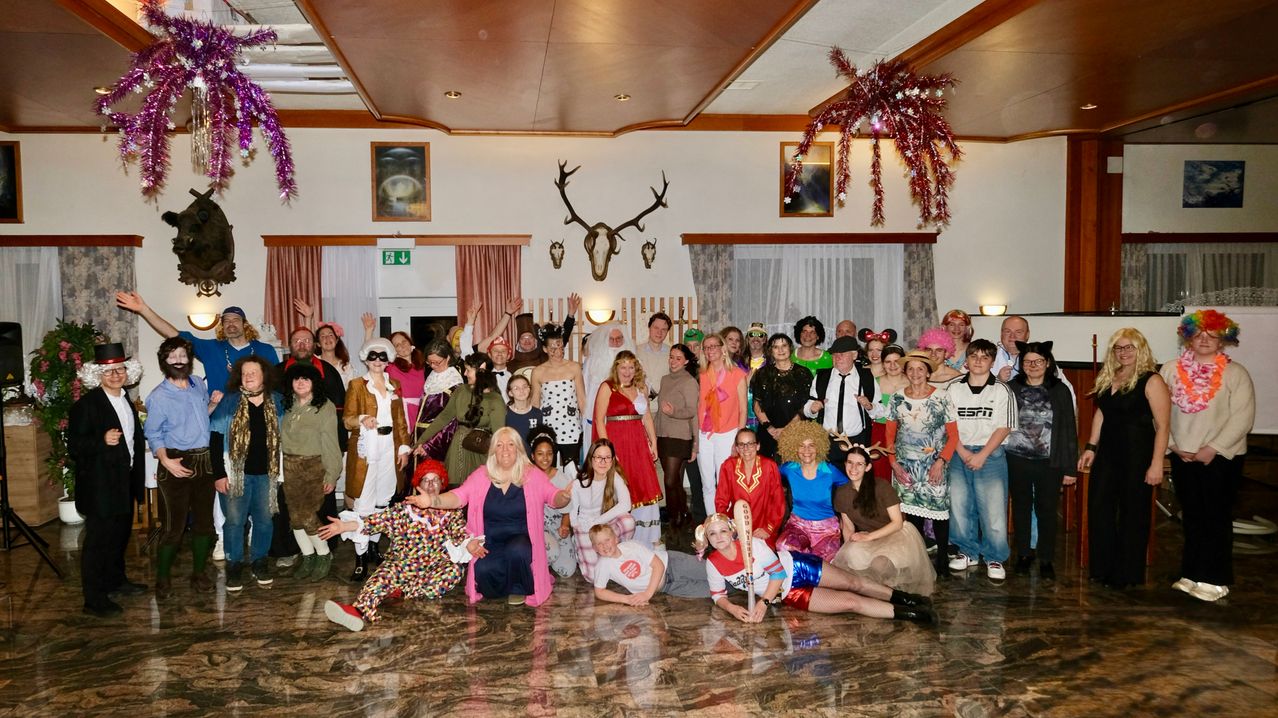 Eine Gruppe von Menschen in verschiedenen Kostümen posiert für ein Foto in einem Raum mit Marmorboden, Hirschköpfen und Weihnachtsdekorationen.