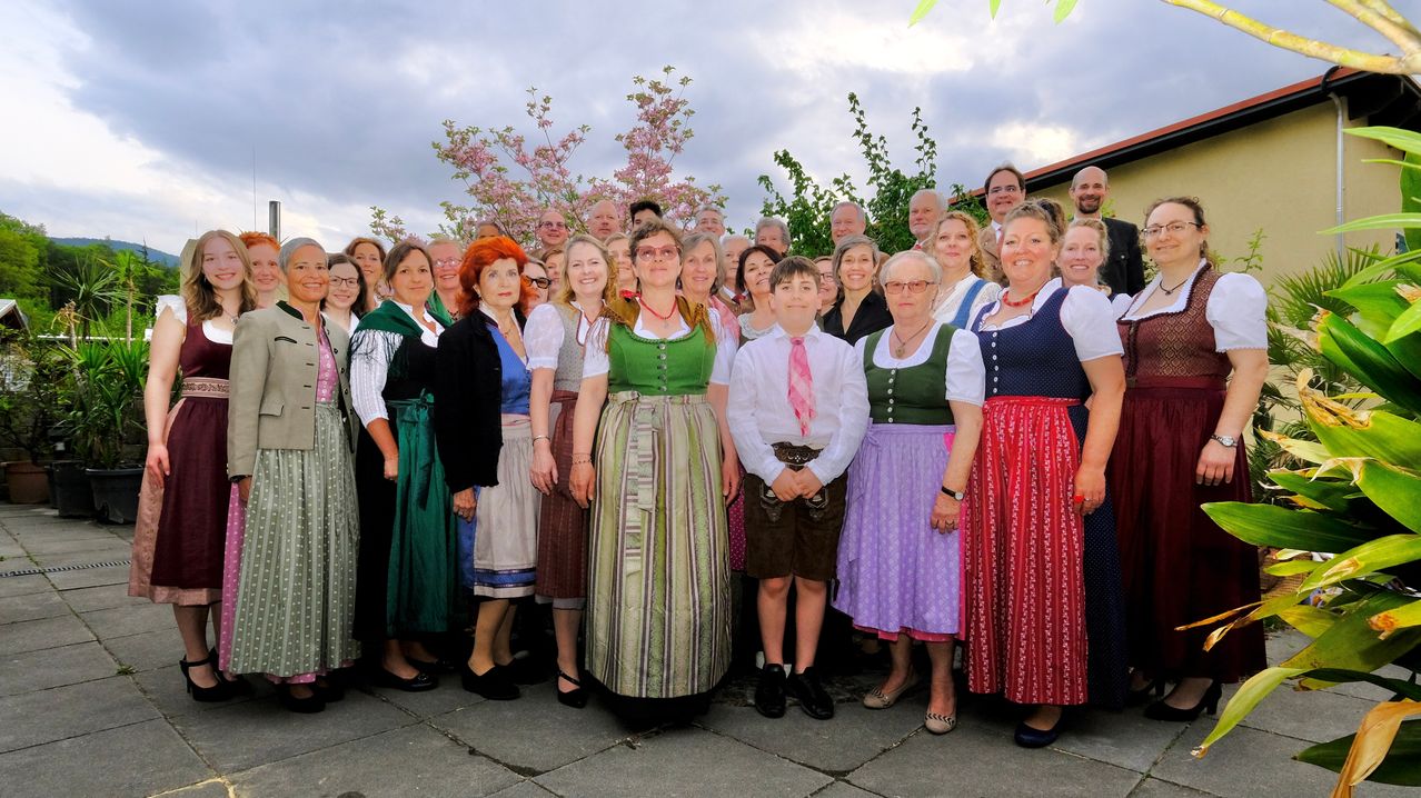 A group of people in traditional clothing stands outside, with trees and a cloudy sky in the background. They are smiling and posing for a photo.