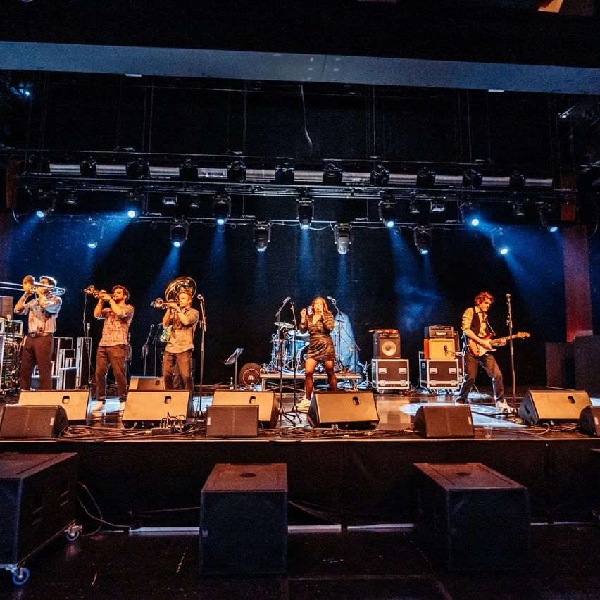 A band performs on stage with a mix of instruments, including trumpets and guitars. The stage is illuminated by blue lights, and speakers are set up in the foreground.