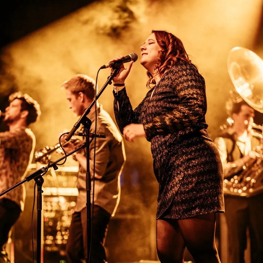 A woman sings passionately on stage with a band, holding a microphone. Behind her, musicians play trumpets and other instruments. The stage is illuminated with warm lighting.