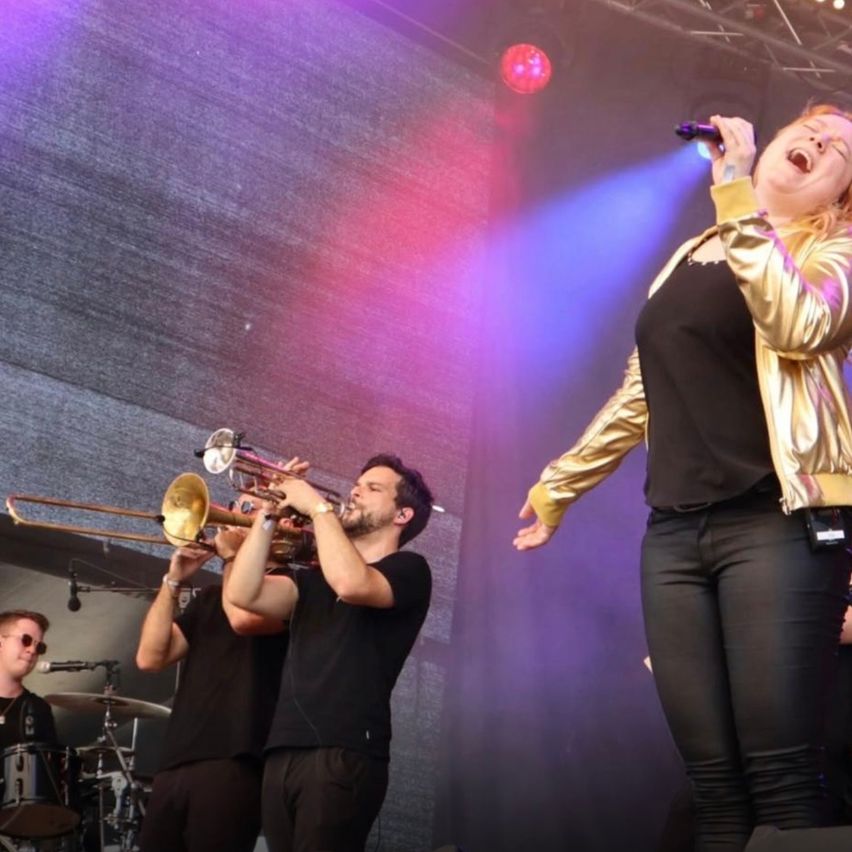 A woman is singing on a stage with a band. She is holding a microphone and wearing a gold jacket. Behind her, two men are playing the trombone and cymbals.