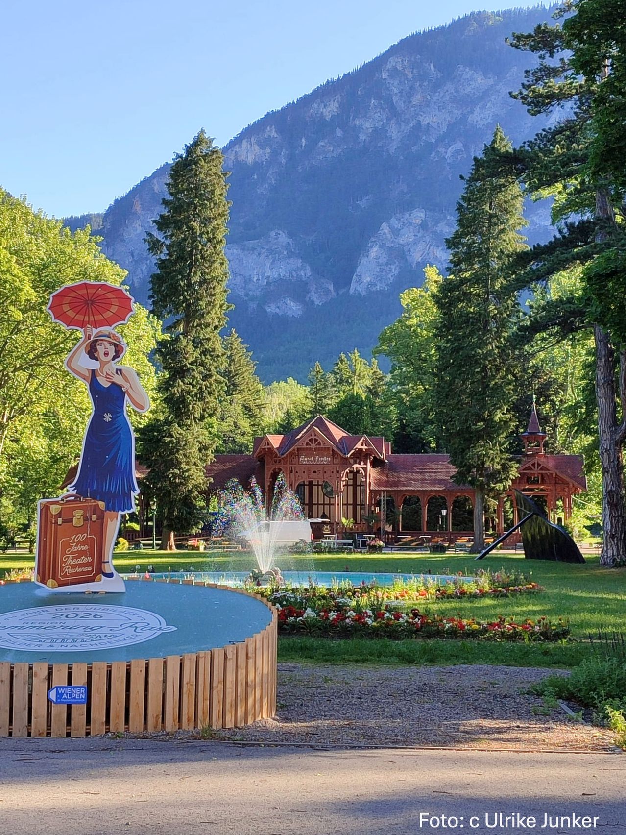 A woman in a blue dress holding an umbrella stands in front of a mountain. Behind her, a fountain and a building with a tower are visible. Trees and flowers adorn the area.