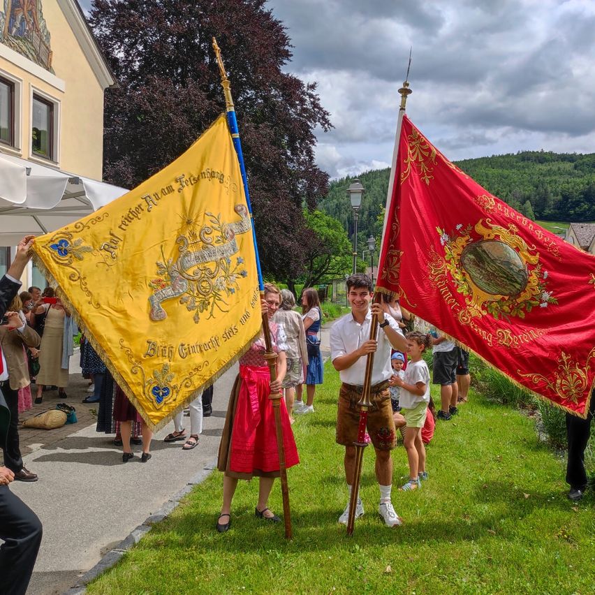 A man and woman hold a yellow and red flag in front of a crowd on a grassy lawn. The flags have intricate designs. The sky is cloudy.