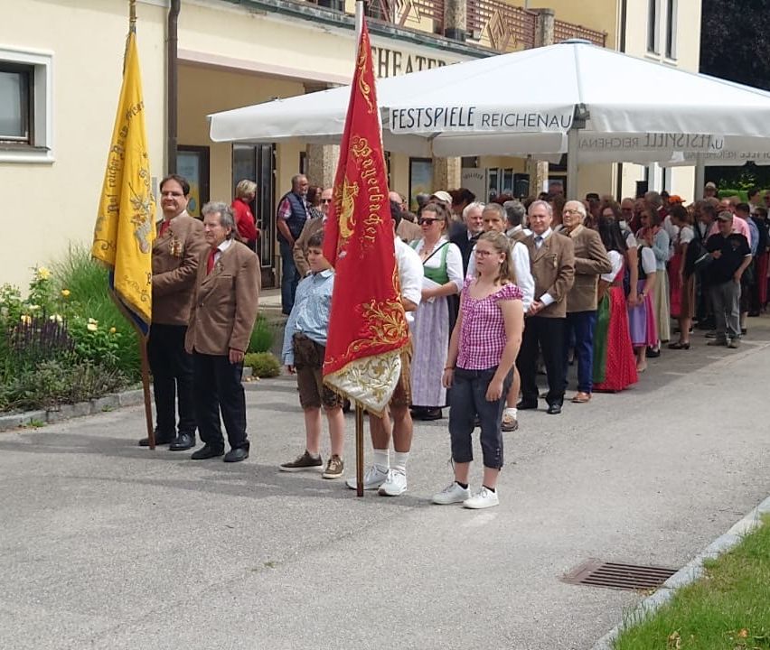 Several people stand in a line in front of a building with a flagpole. They wear traditional clothes and hold flags. A white tent with the word 'FESTSPIELE REICHENAU' is behind them.