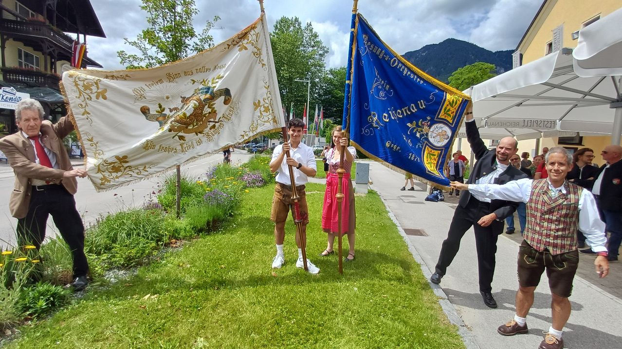 Two people in traditional clothing hold flags on a grassy path. A third person stands behind them, and a mountain is in the distance.