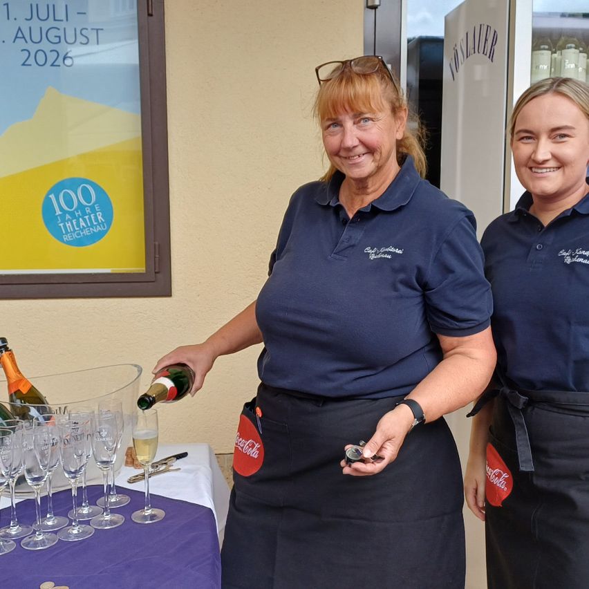 Two women in blue polo shirts and black aprons are preparing drinks. One is pouring champagne, while the other holds a corkscrew. Behind them, a poster advertises the theater.