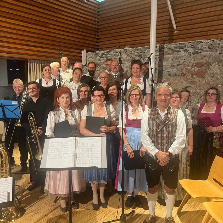 A group of musicians in traditional attire pose for a photo with music stands and instruments in a rehearsal space.