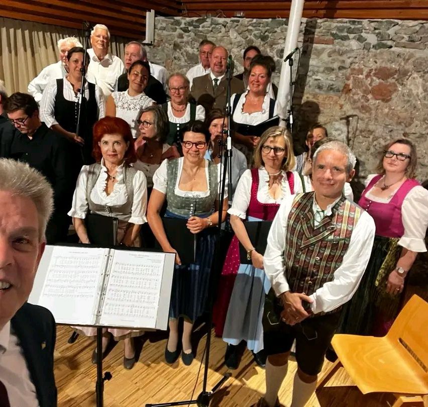 A choir dressed in traditional clothing stands with a music stand in front of a microphone. The background shows stone walls and a curtain.