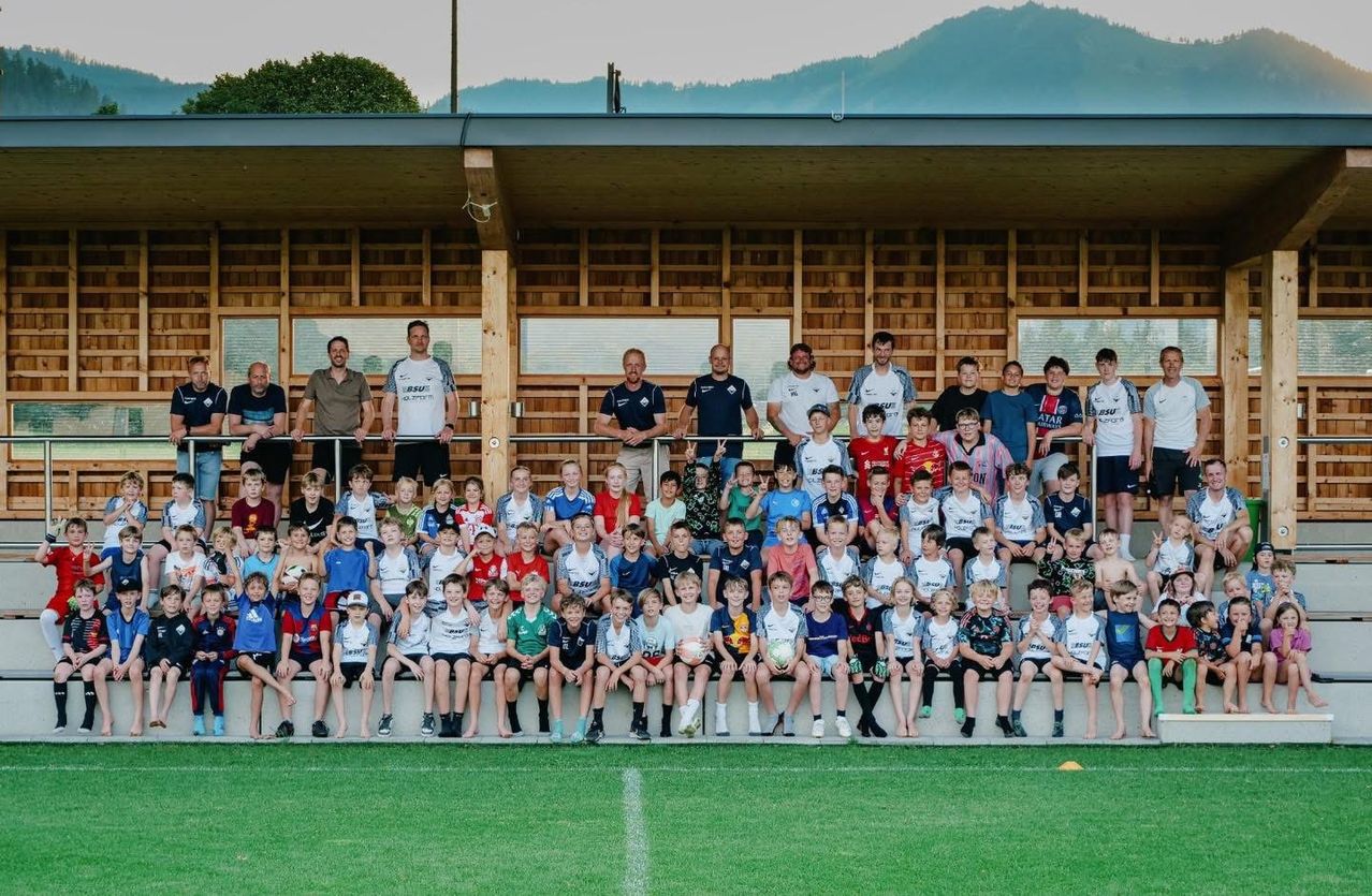 Eine Gruppe von Kindern und Erwachsenen posiert für ein Foto auf einem Sportfeld mit einem Holzgebäude und Bergen im Hintergrund.