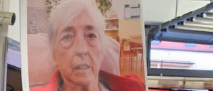 Eine Person hält ein Foto einer älteren Frau in einem roten Mantel und einem lila Hemd. Das Foto befindet sich auf einem roten Auto.