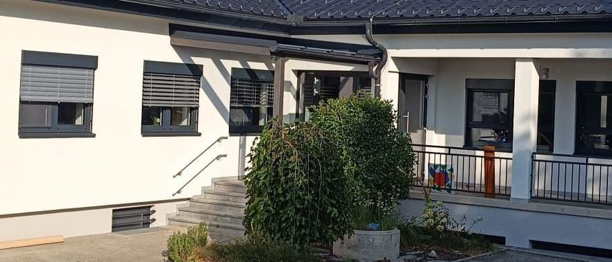 Ein weißes Haus mit schwarzem Dach und Fenstern mit Jalousien. Eine Treppe führt zu einer Veranda mit einem Geländer. Eine Pflanze steht an der Seite.