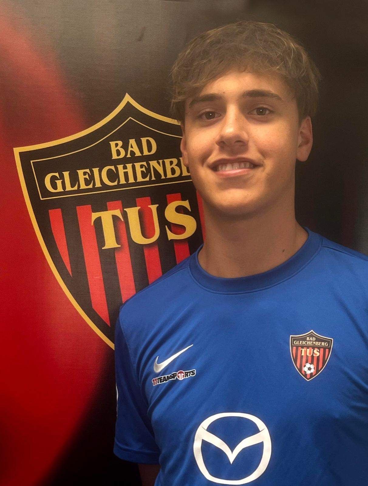 Ein junger Mann in einem blauen Fußballtrikot lächelt vor einem rot-schwarzen Sportteambanner.