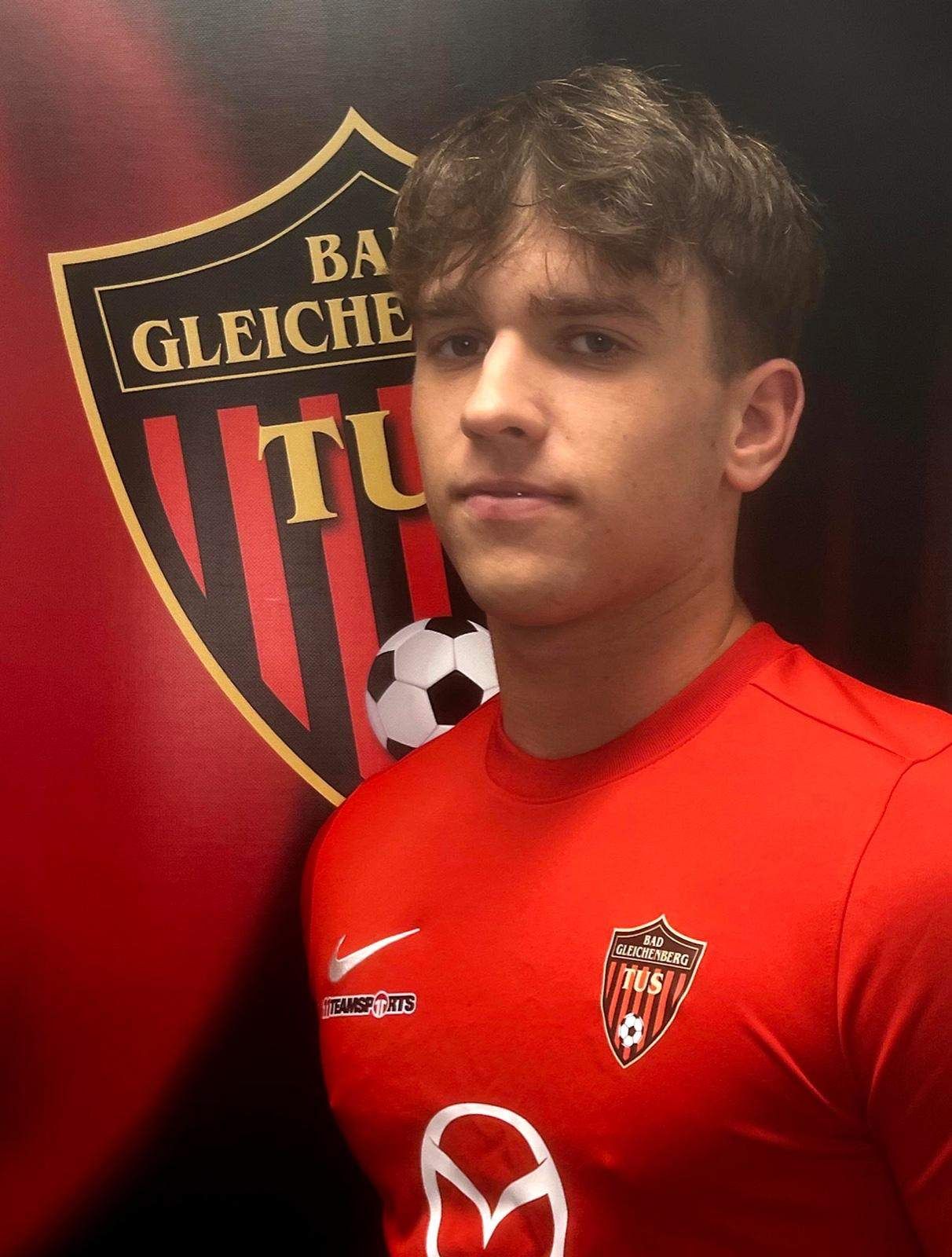 Ein junger Mann steht vor einer roten Wand mit einem Fußballlogo und trägt ein rotes Fußballtrikot mit einem Nike-Logo auf der linken Seite.