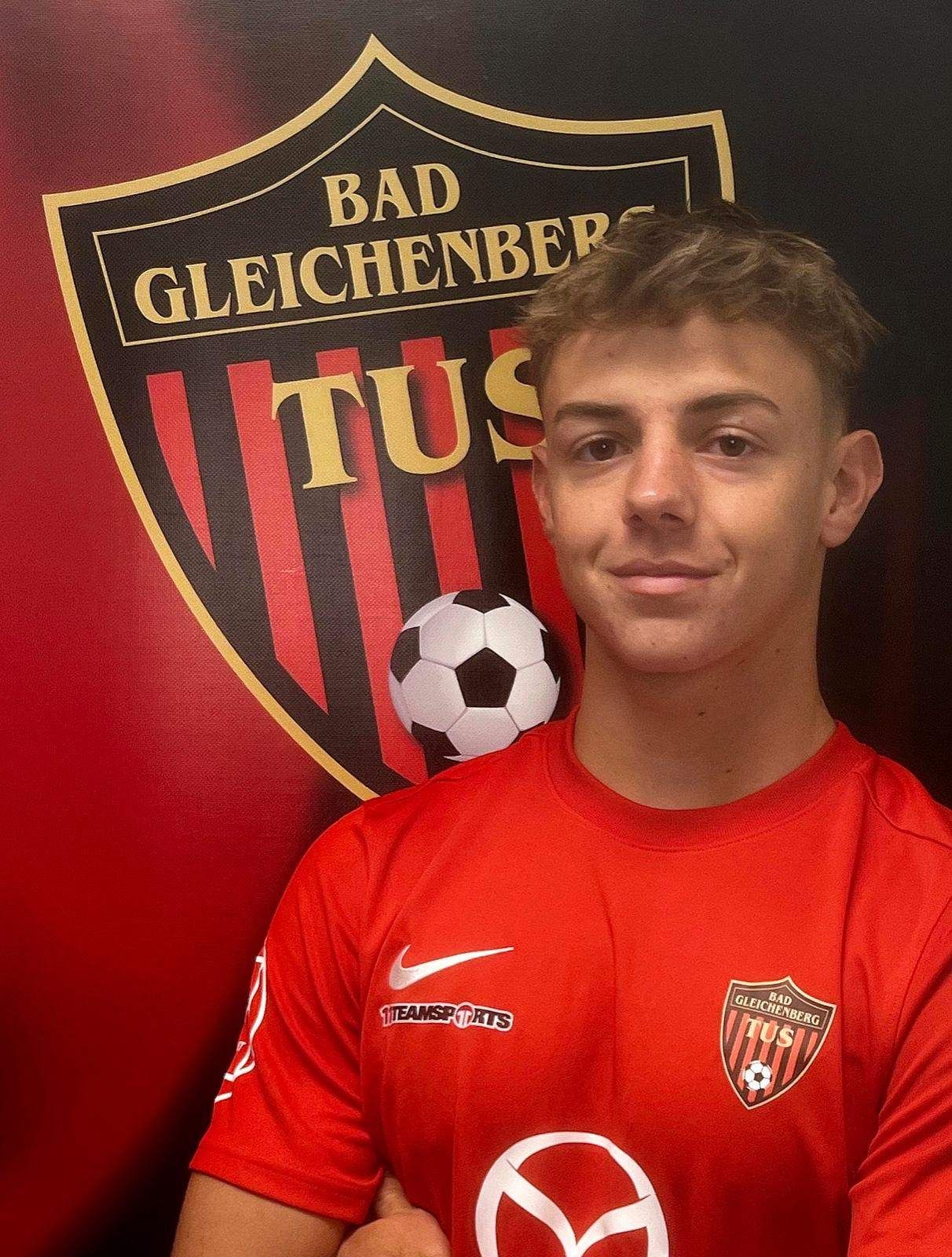 Ein Mann in einem roten Fußballtrikot mit einem Fußball auf der Schulter posiert vor einer roten Wand mit dem Logo von Bad Gleichenberg.