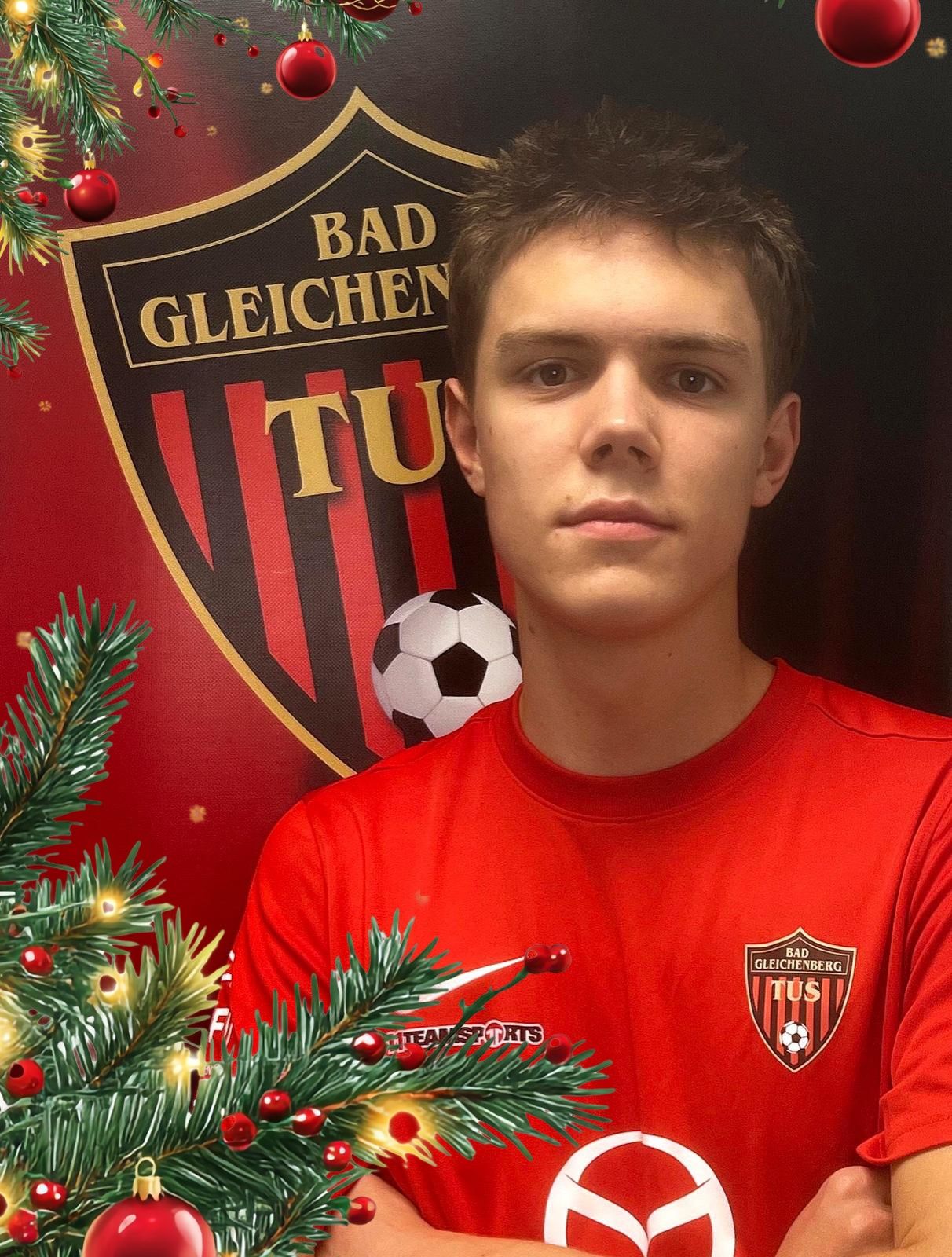 Ein junger Mann in einem roten Shirt posiert für ein Foto vor einem Weihnachtsbaum und einem Fußball, mit einem roten und schwarzen Fußballteam-Emblem im Hintergrund.