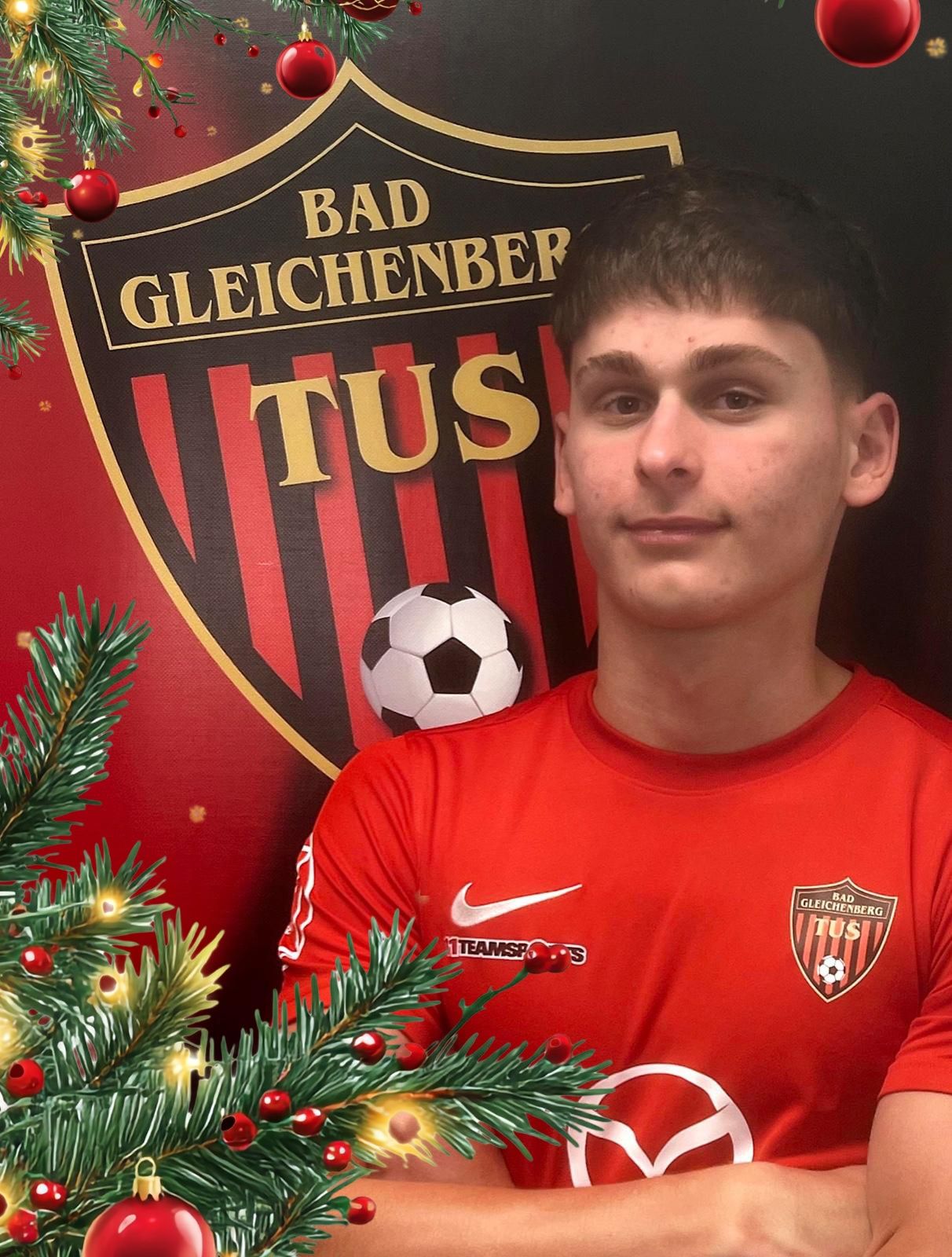 Ein junger Mann posiert in einem roten Fußballtrikot vor einem Weihnachtsbaum und einem Fußball. Hinter ihm ist das Logo des Fußballvereins Bad Gleichenberg TSU zu sehen.