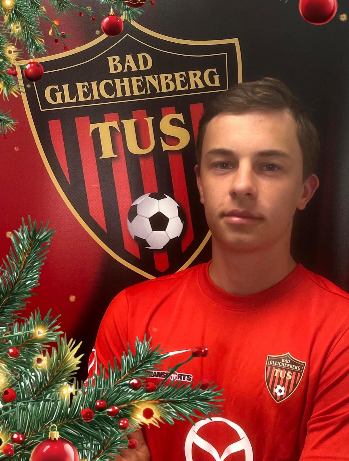 Ein Mann in einem roten Fußballtrikot posiert vor einem Bad Gleichenberg Logo, einem Weihnachtsbaum und einem Fußball.