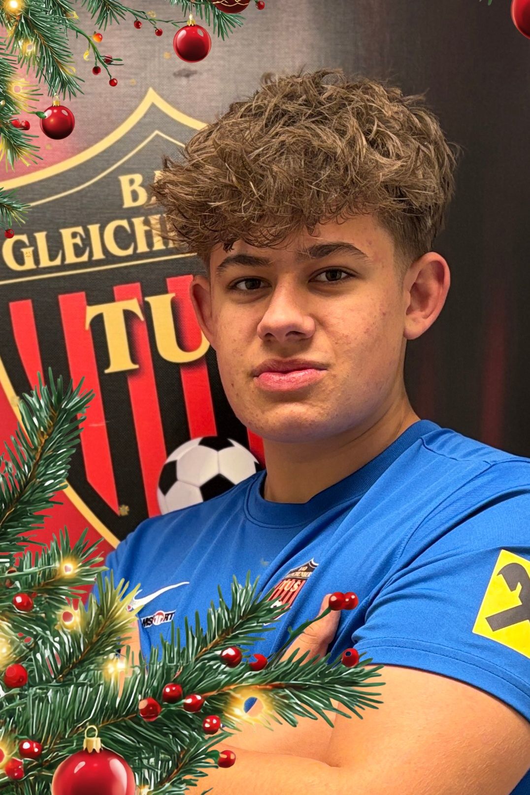 Ein junger Mann posiert mit einem Weihnachtsbaum vor dem Logo einer Fußballmannschaft. Er trägt ein blaues Trikot mit der Nummer 3 am Ärmel.
