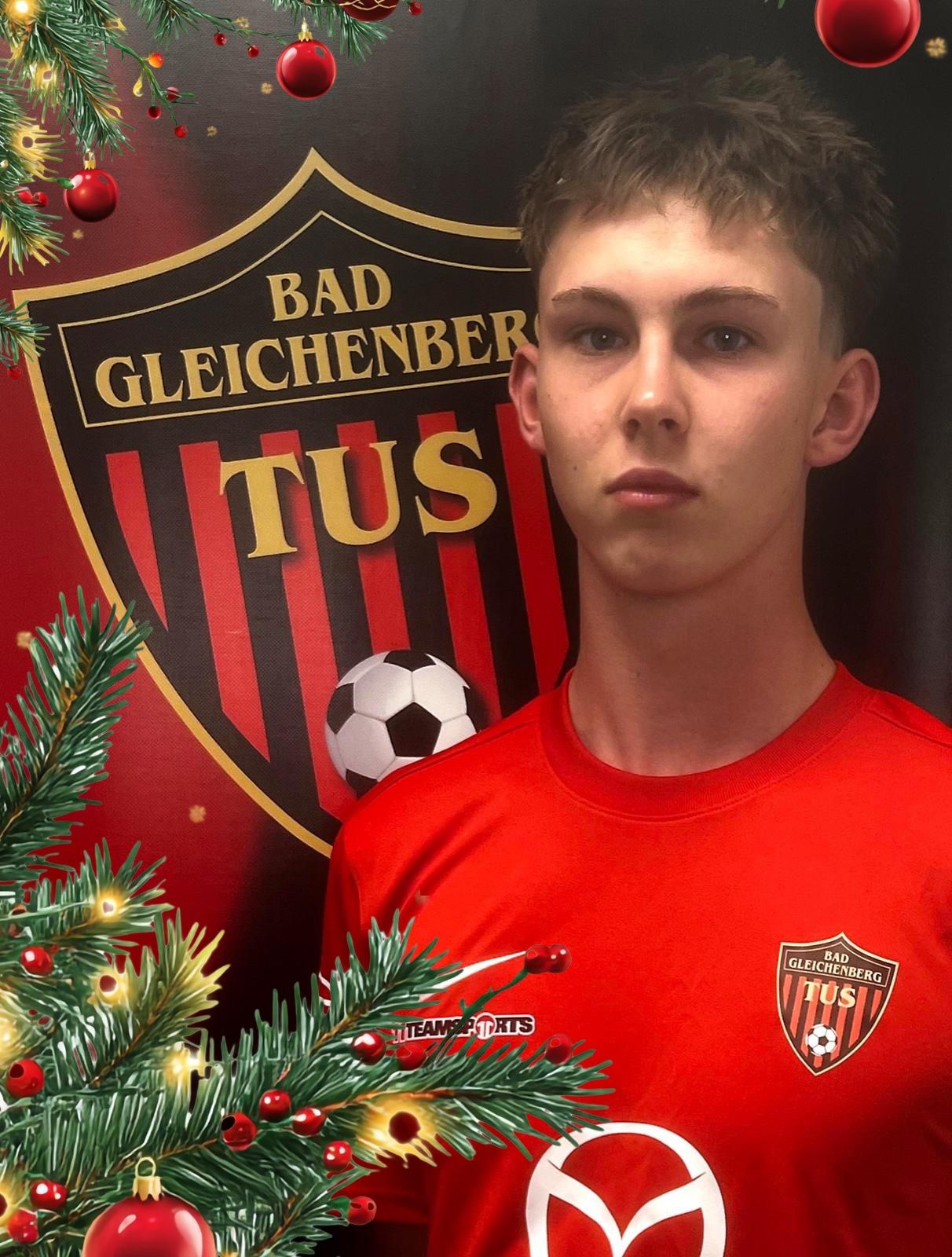 Ein junger Mann in einem roten Fußballtrikot steht vor einem festlichen Hintergrund, der einen Weihnachtsbaum und das Logo des Fußballvereins Bad Gleichenberg Tus zeigt.
