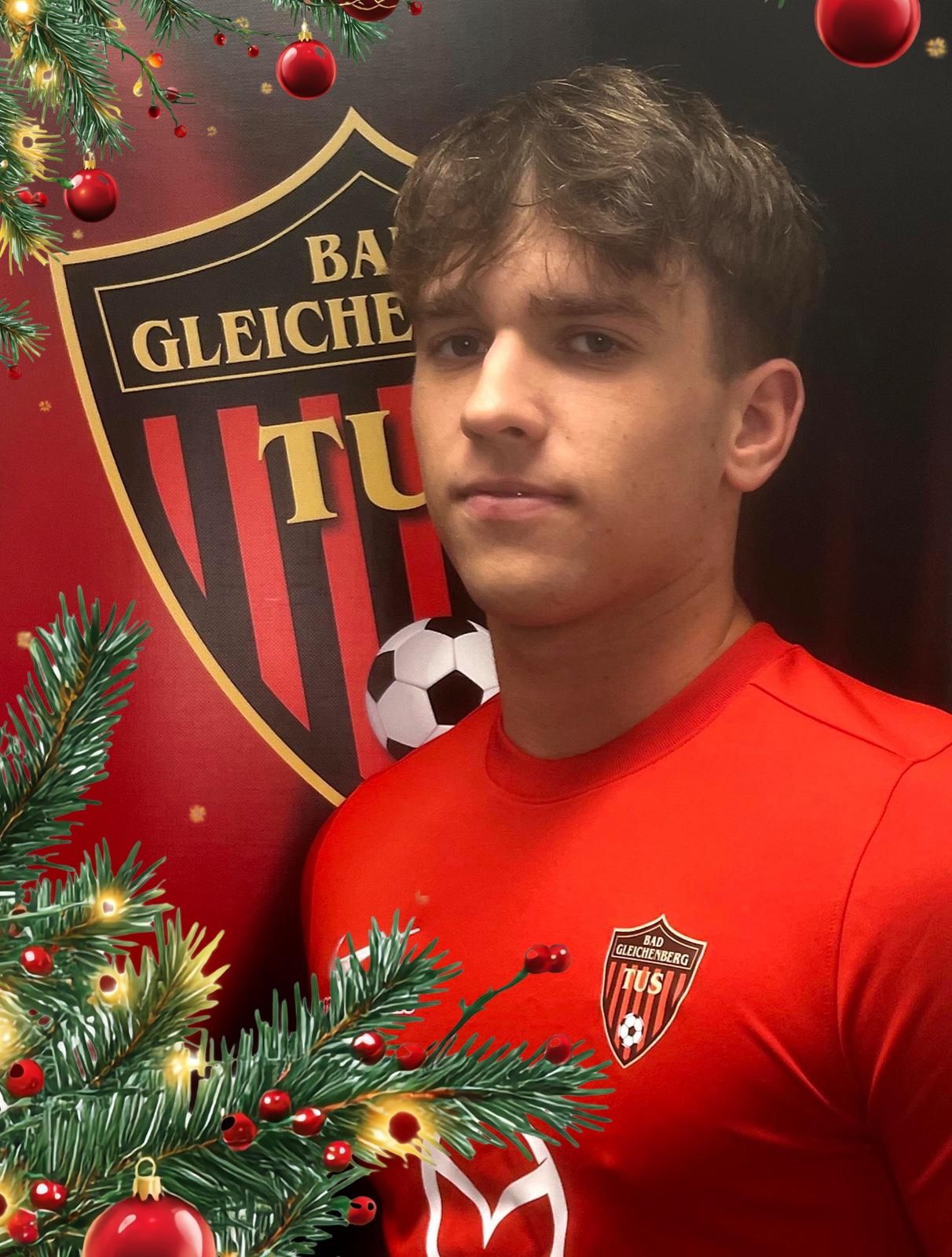 Ein Mann in einem roten Fußballtrikot steht vor einem Weihnachtsbaum und einem Fußball. Der Hintergrund zeigt ein rotes Logo mit der Aufschrift 'BA GLEICHE TU'.