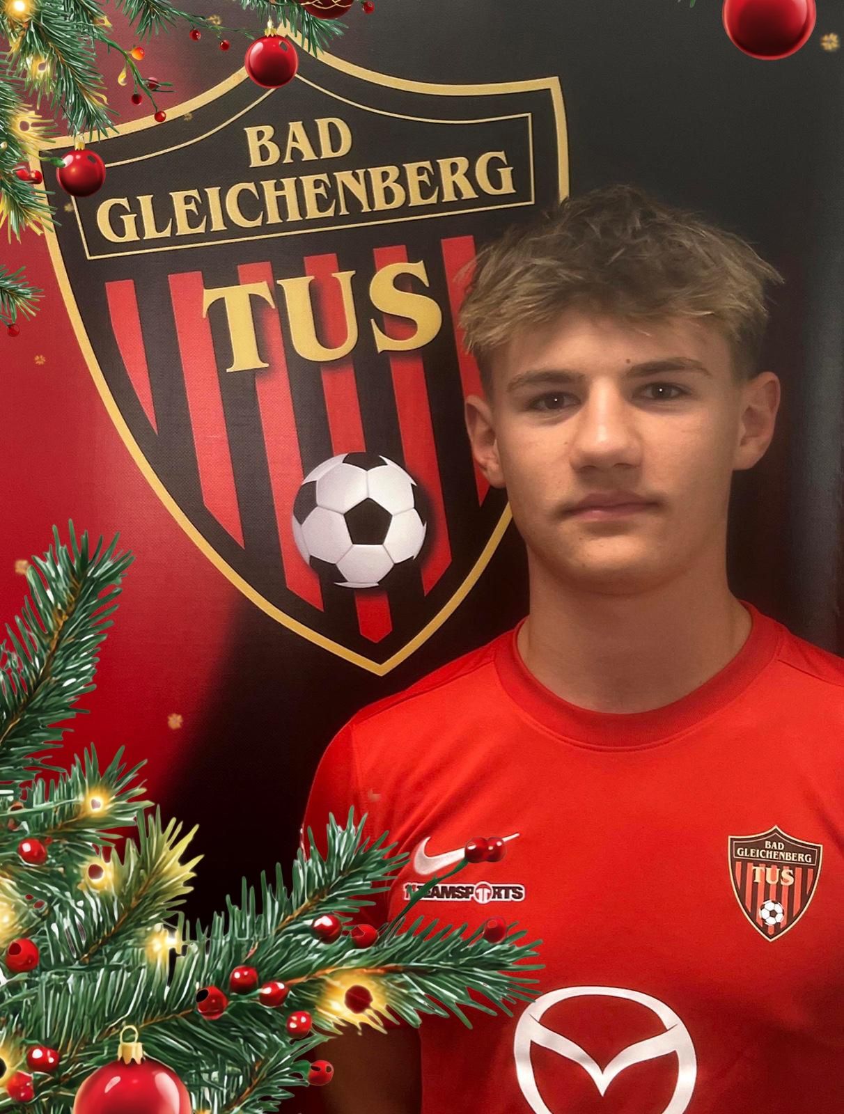 Ein junger Mann in einem roten Fußballtrikot steht vor einem rot-schwarzen Teamlogo, einem Fußball und einem Weihnachtsbaum.