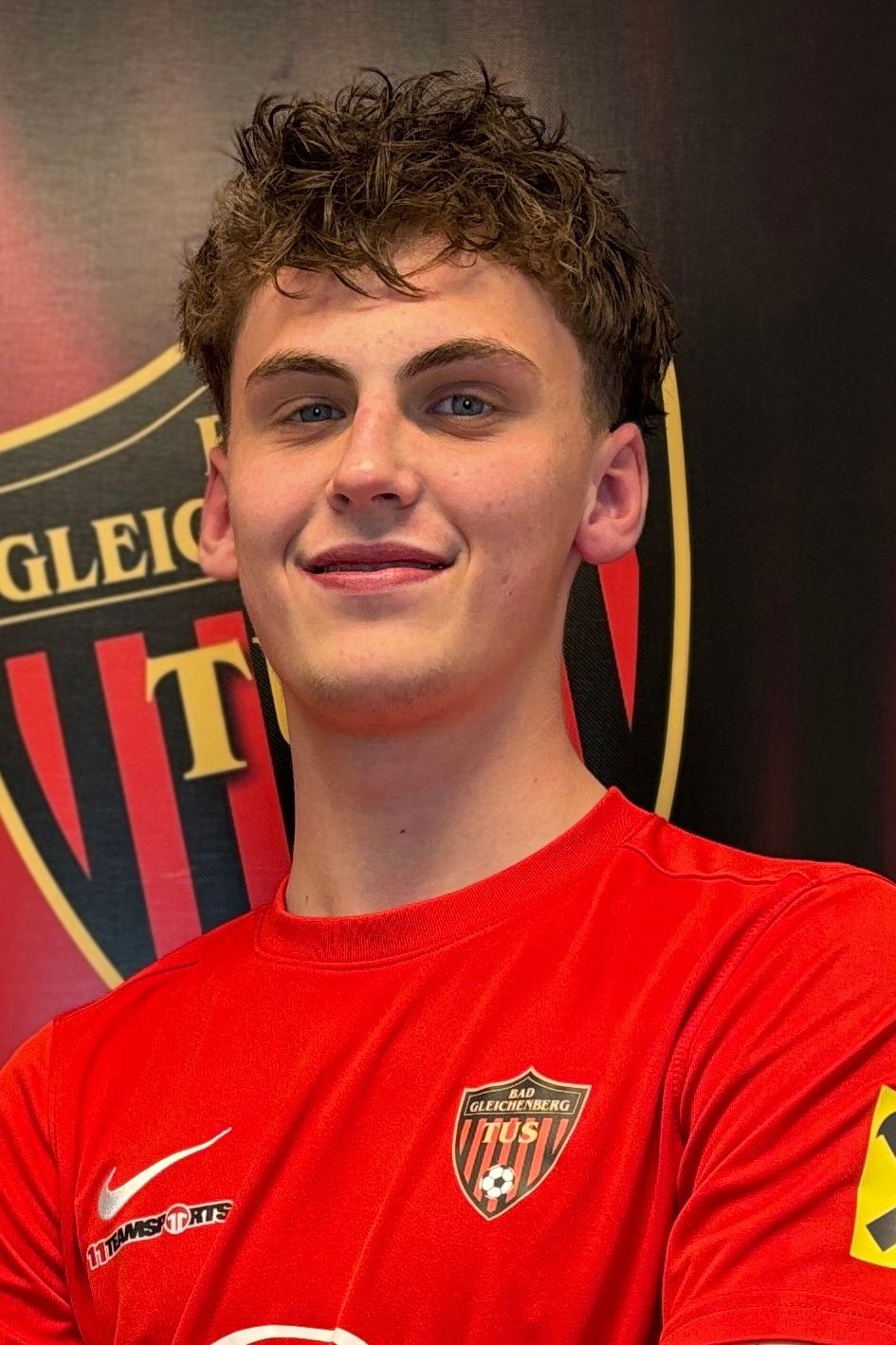 Ein Mann steht vor einer Wand mit einem großen Logo und trägt ein rotes T-Shirt mit Logo auf der linken Seite.