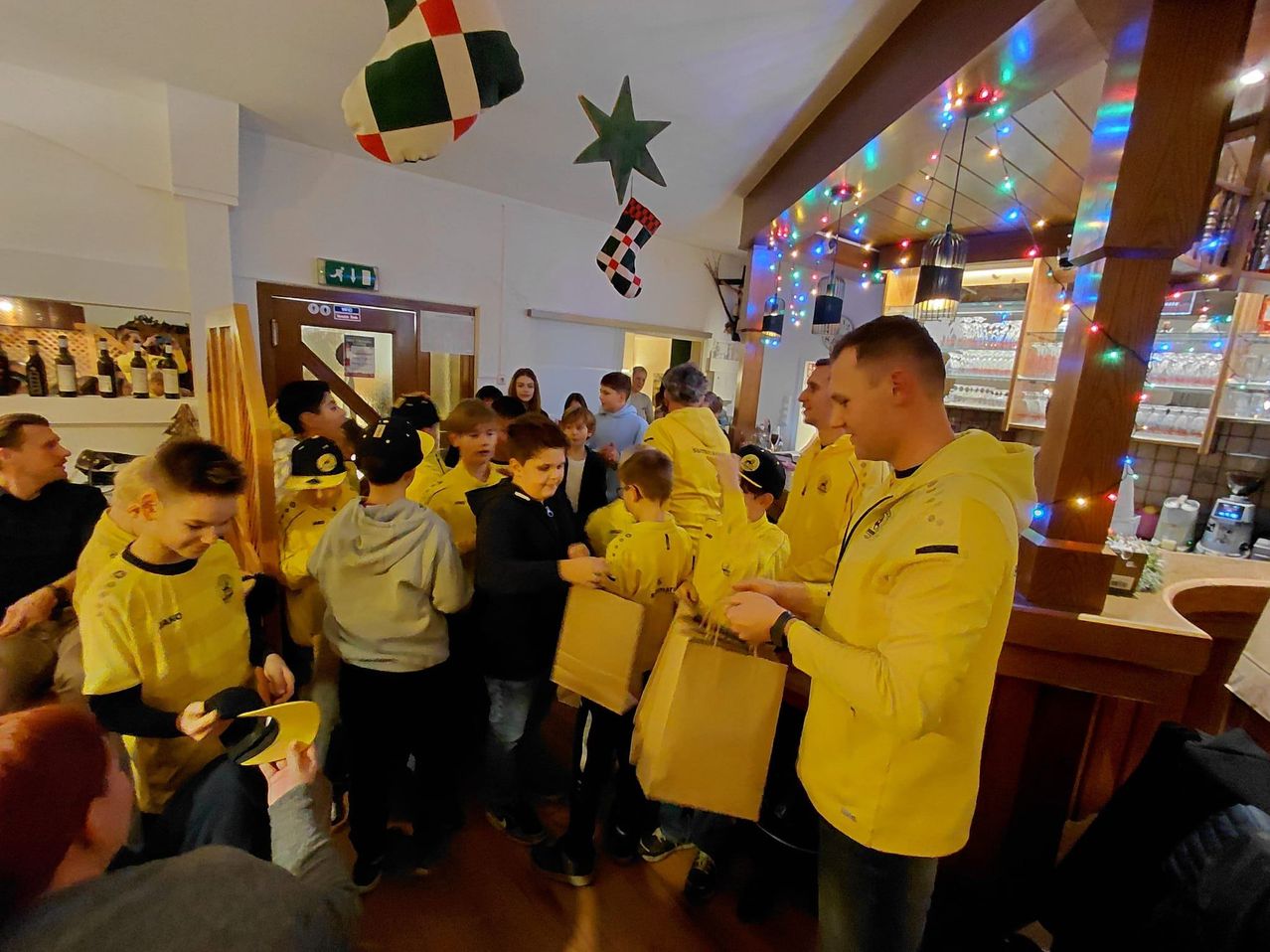 Eine Gruppe von Kindern und Erwachsenen versammelt sich in einem festlich geschmückten Raum und erhält Geschenke in Papiertüten. Sie sind in gelben Jacken und Mützen gekleidet. Ein Mann verteilt die Geschenke. Der Raum ist mit Weihnachtslichtern und Ornamenten geschmückt.