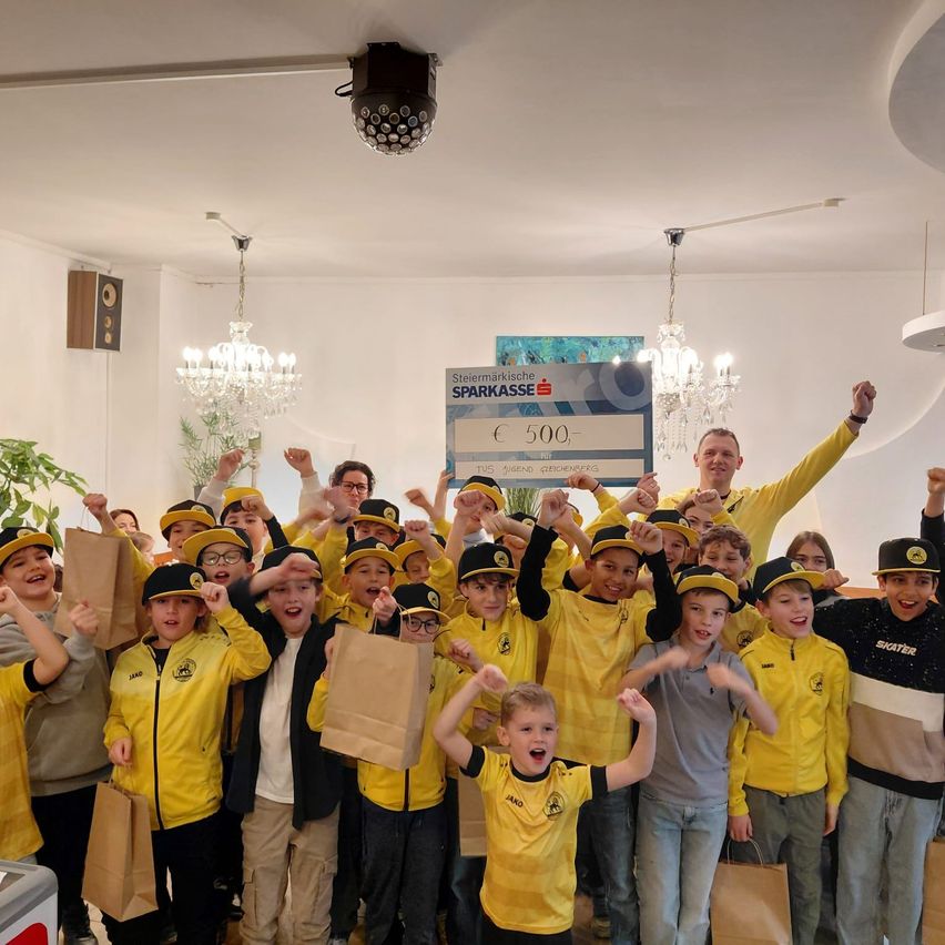 Eine Gruppe von Kindern in gelben Trikots und Mützen feiert mit einem großen Scheck und Taschen. Ein Mann hält einen Scheck mit dem Text '500', und die Kinder heben ihre Hände.