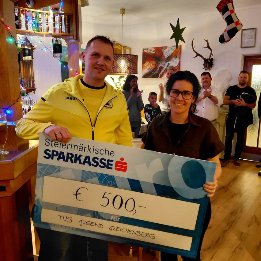 Ein Mann und eine Frau halten einen großen Scheck über 500 Euro. Sie lächeln und posieren für ein Foto in einem dekorierten Raum. Hinter ihnen klatschen Menschen und stehen in der Nähe eines Kamins.