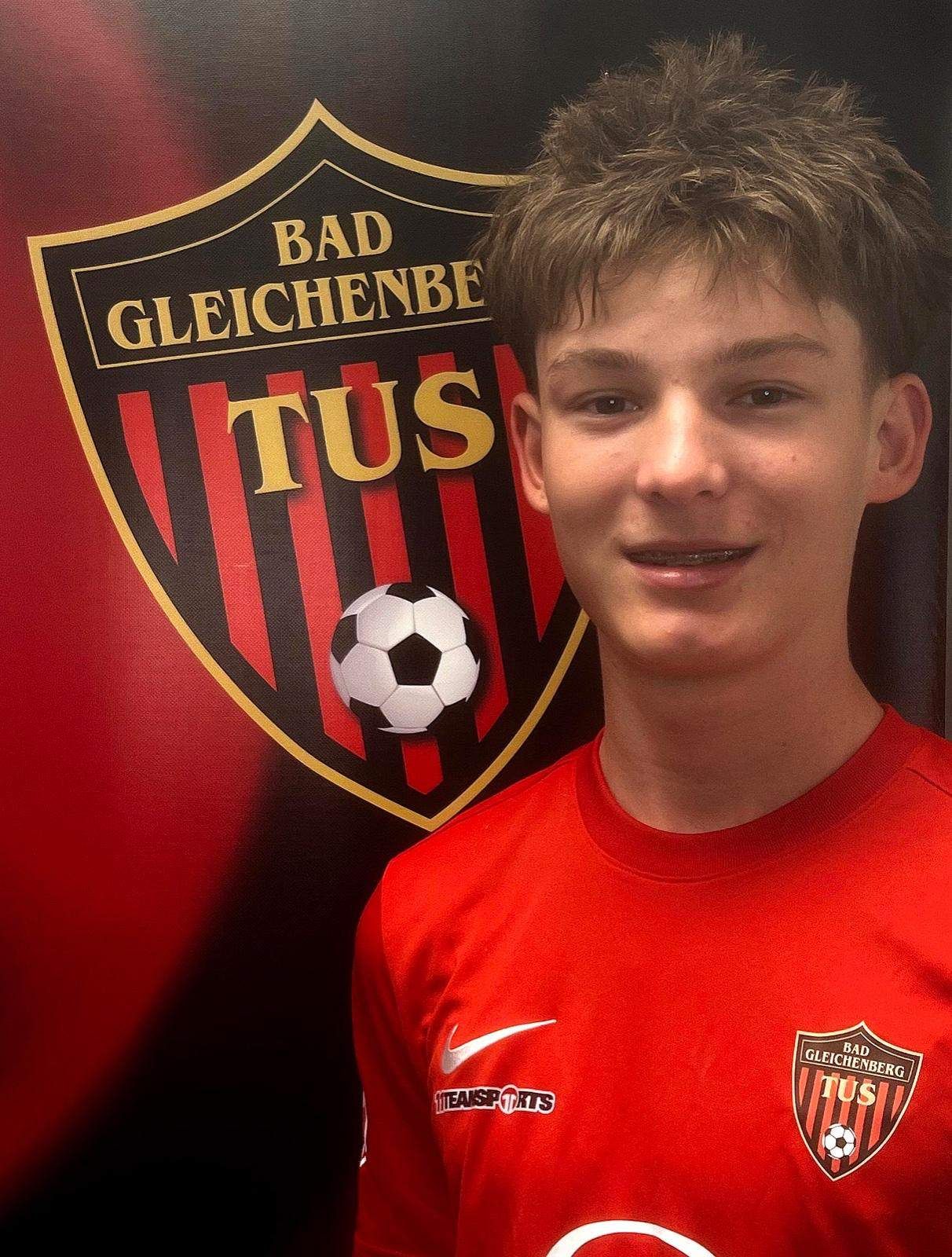 Ein junger Mann in einem roten Shirt steht vor einem Fußballvereinslogo. Das Logo ist rot und schwarz mit den Buchstaben 'TuS' in weiß. Er hat einen Fußball auf der Schulter.