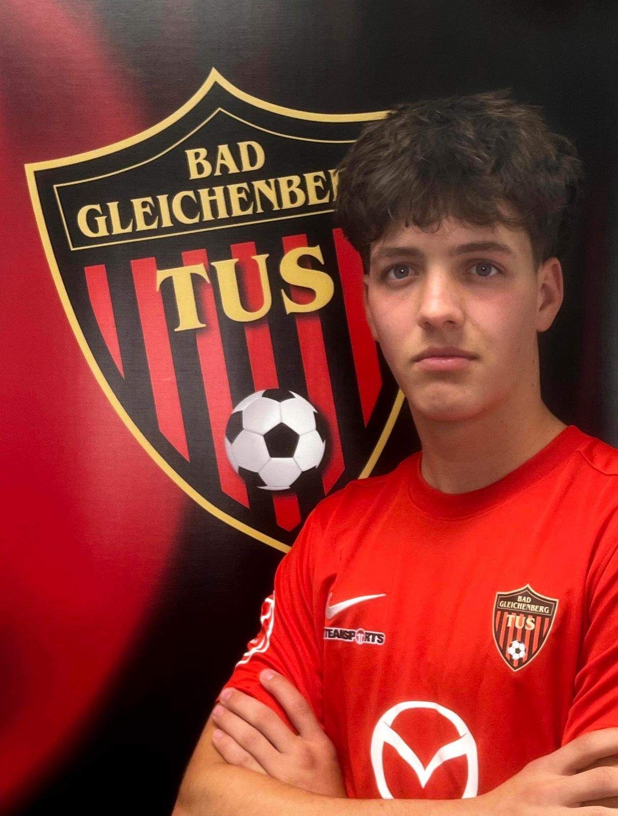 A young man wearing a red soccer jersey with the logo of Bad Gleichenberg stands in front of a soccer ball floating in the air.