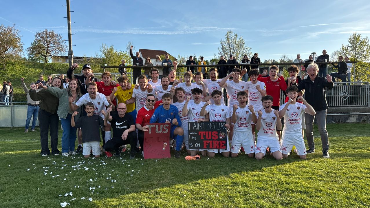 Eine Fußballmannschaft feiert auf dem Spielfeld mit Fans im Hintergrund. Ein Spieler hält ein Schild, auf dem steht 'ICH BIN KEIN LÜGNER, wenn du sagst, TUS BRENNT'.