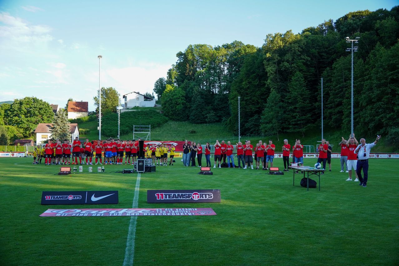 Eine Gruppe von Menschen in roten und gelben Uniformen steht auf einem Feld. Sie klatschen und jubeln. Es gibt Sponsorenbanner auf dem Feld. Bäume und Häuser sind in der Ferne.