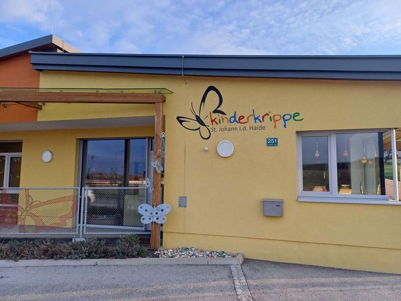 Ein gelbes Gebäude mit dem Namen Kinderkrippe St. Johann i.d. Haide, versehen mit einem Schmetterlingsdesign. Der Eingang hat einen Zaun, und es gibt zwei Fenster mit einem Briefkasten auf der rechten Seite.