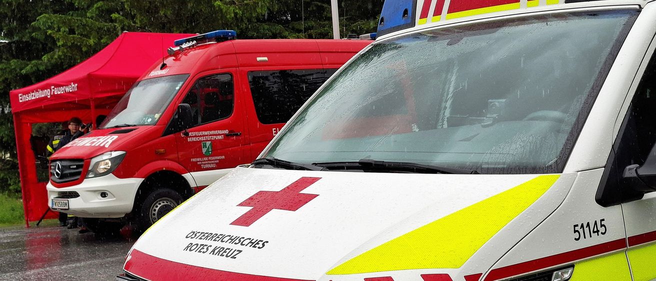 Zwei Ambulanzfahrzeuge, beide mit rotem Kreuz und gelben Reflektoren markiert, sind bei Regen geparkt. Das vordere Fahrzeug hat 'Osterreichisches Rotes Kreuz' auf seiner Motorhaube.