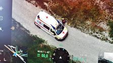 Ein weißer Krankenwagen ist auf einem Kiesweg geparkt. Eine Person in einem gelben Helm steht neben dem Fahrzeug. Das Bild zeigt einen Tachometer mit 00,0 m/s und eine Kompassanzeige mit einer Höhe von 187,4 m.