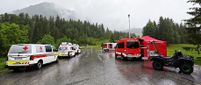 Mehrere Rettungsfahrzeuge sind auf einer nassen Straße mit Berghintergrund geparkt. Ein Fahrzeug hat 'Feuerwehr' darauf geschrieben.
