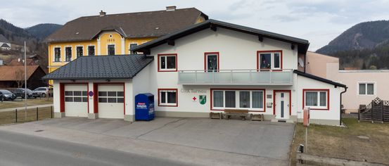 Ein weißes Gebäude mit roten Akzenten und einem Balkon trägt das Schild 'Orts Turnau'. Es hat einen blauen Briefkasten, zwei Garagen und eine Bank davor. Ein gelbes Gebäude mit braunem Dach steht dahinter.