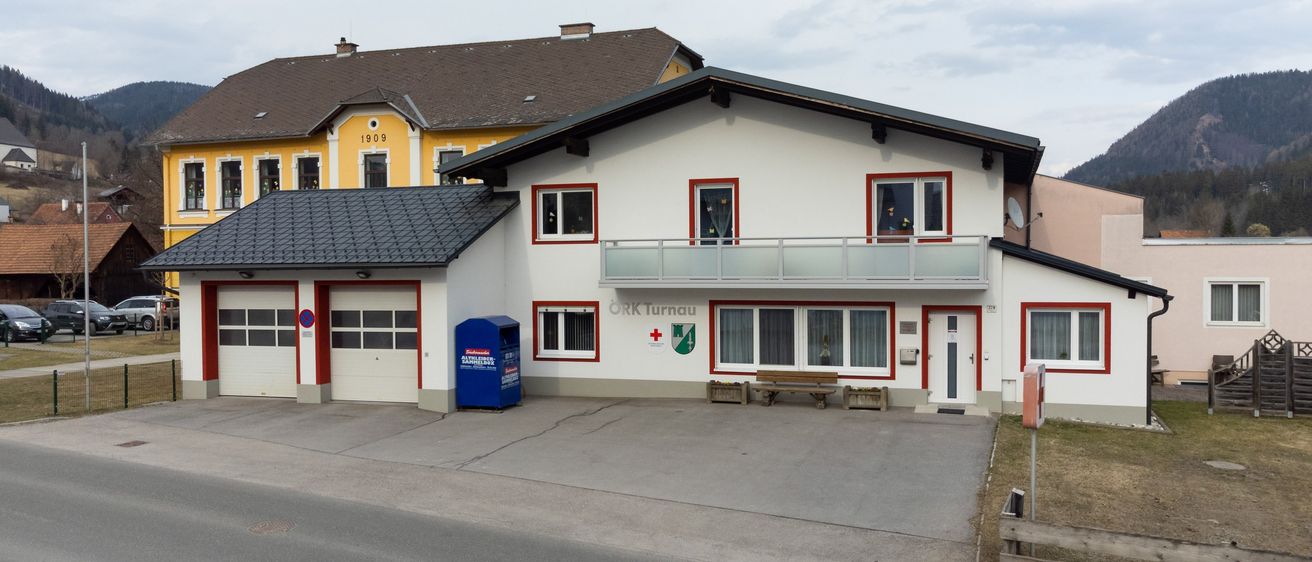 Ein weißes Gebäude mit roten Akzenten und einem Balkon trägt das Schild 'Orts Turnau'. Es hat einen blauen Briefkasten, zwei Garagen und eine Bank davor. Ein gelbes Gebäude mit braunem Dach steht dahinter.