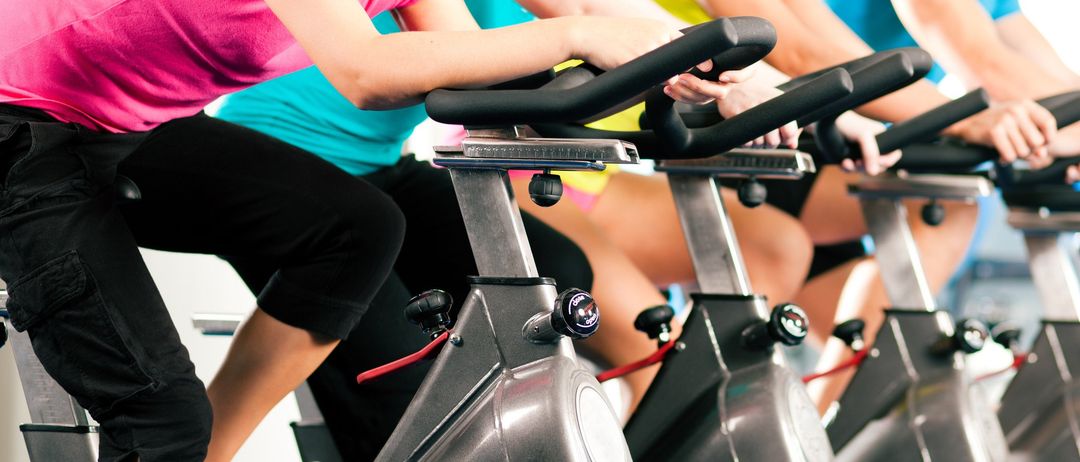 A group of women in workout gear are riding spinning bikes in a gym. They are all wearing sneakers and some have bracelets.