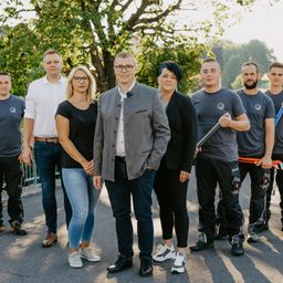 Eine Gruppe von Menschen steht in einer Reihe draußen. Sie sind in verschiedenen Outfits gekleidet, und einer hält ein Werkzeug. Im Hintergrund sind Bäume und ein Gebäude zu sehen.