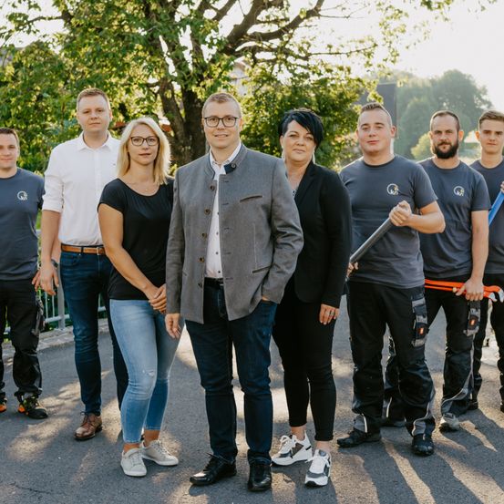 Eine Gruppe von Menschen steht in einer Reihe draußen. Sie sind in verschiedenen Outfits gekleidet, und einer hält ein Werkzeug. Im Hintergrund sind Bäume und ein Gebäude zu sehen.