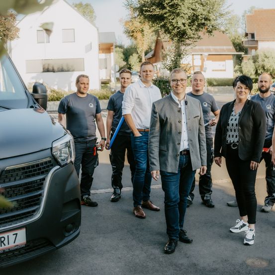 Eine Gruppe von Menschen steht auf einem Parkplatz vor einem Auto und einem Haus. Sie tragen Freizeitkleidung und einige halten Werkzeuge. Sie scheinen für ein Foto zu posieren.