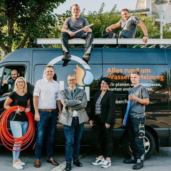 Eine Gruppe von Menschen, einige stehend und einige auf einem Van sitzend, posieren für ein Foto. Der Van hat ein Logo und Text auf seiner Seite.