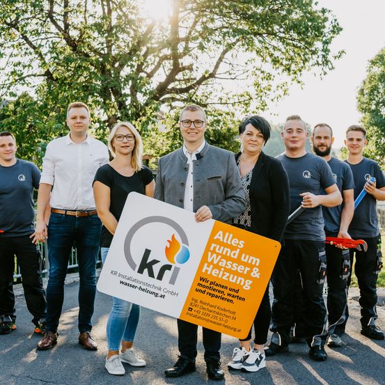 Eine Gruppe von Personen steht im Freien und hält ein Schild mit der Aufschrift 'KRI Installationstechnik GmbH Wasser-Heizung.at'. Sie scheinen Klempner oder Techniker zu sein, die lächelnd für ein Foto posieren.