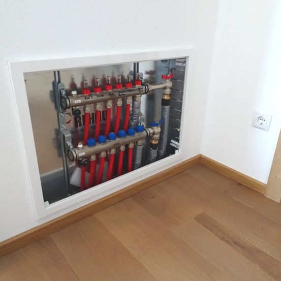 Eine Ecke eines Raumes mit weißer Wand, Holzboden und einer weiß gerahmten Metallplatte, die ein komplexes Heizsystem mit verschiedenen Rohren und Ventilen zeigt.