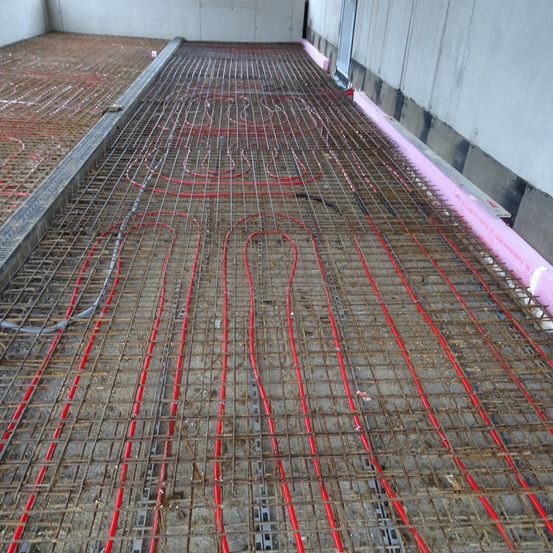 Ein Innenausbau mit einem Betonboden und roten Rohren, die auf einem Metallgitter verlegt sind. Die Wände sind weißer Beton.