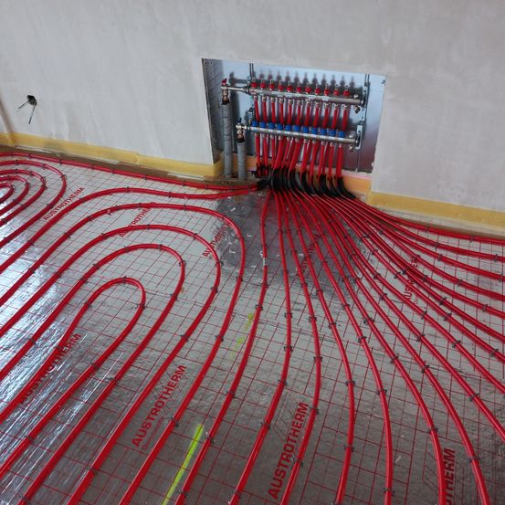 Rote Heizungsrohre sind in einem Rastermuster auf dem Boden verlegt. Die Rohre sind mit einer großen Platte an der Wand verbunden, die verschiedene Rohre und Ventile aufweist.