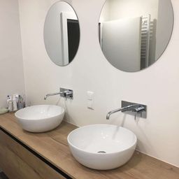 Ein Badezimmer mit einem Holzarbeitsplatten, zwei ovalen Spiegeln und zwei weißen Waschbecken mit Wasserhähnen. Unten befindet sich ein weißer Schrank und links sind Toilettenartikel.